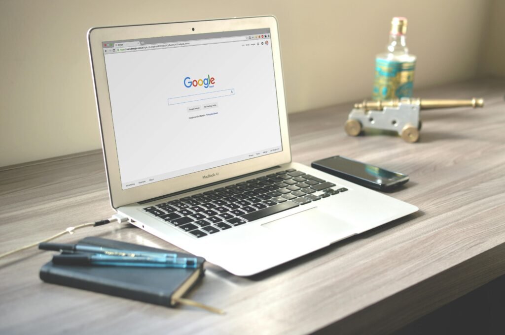 
An open MacBook displaying the Google homepage sits on a wooden desk beside a smartphone, notebook with pens, and a decorative item resembling a miniature cannon.