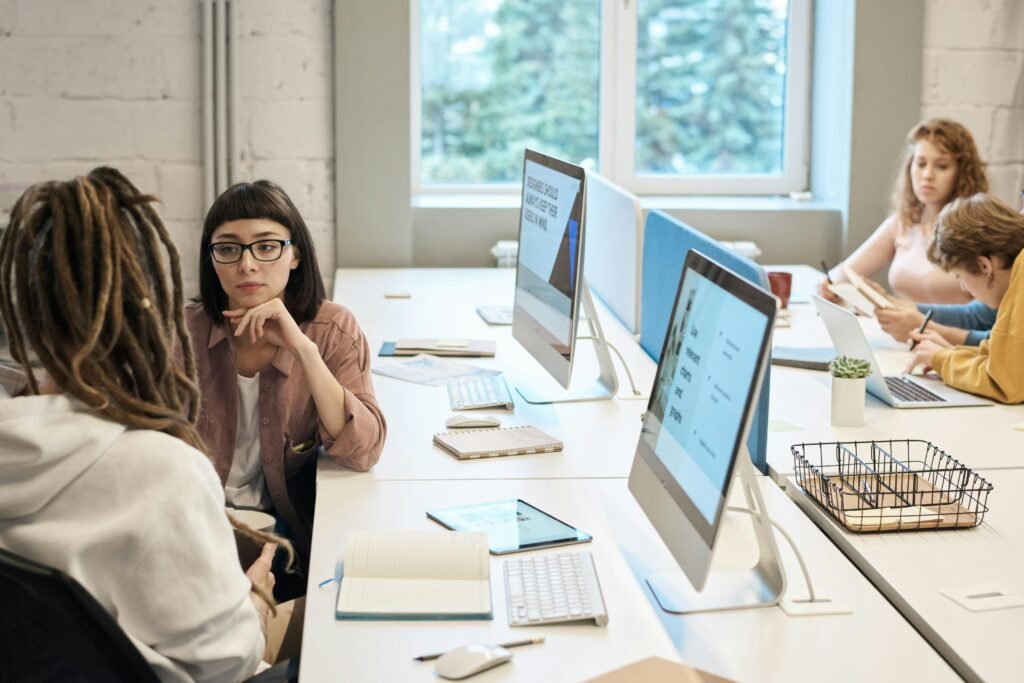 People working and conversing at a shared office table with desktop computers, notebooks, and office supplies, in a modern, naturally lit workspace.