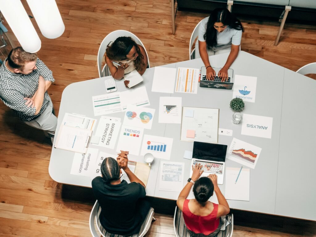 Five people gathered around a large table covered with charts, graphs, and laptops, engaged in a business meeting or team discussion in a modern office space.
