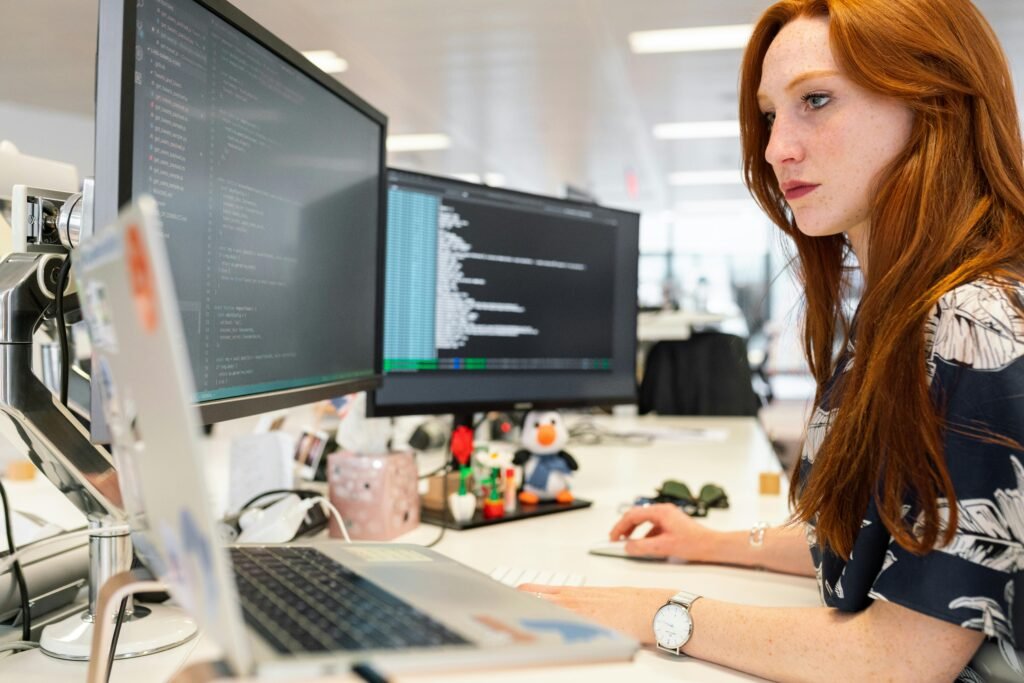 A woman with long red hair works at a desk with multiple monitors displaying code, suggesting software development or programming in a modern office setting.