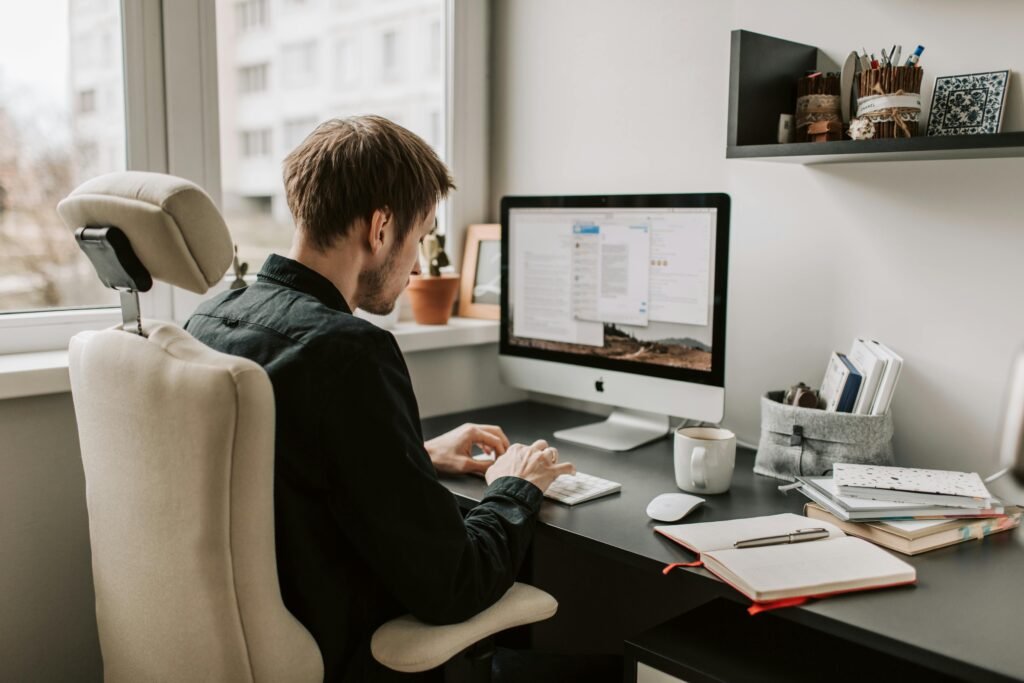 
A man sits at a desk working on a desktop computer in a bright home office, surrounded by notebooks, a coffee mug, and organized shelves with office supplies.
